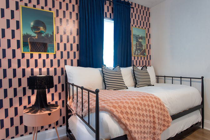 Retro-chic bedroom with black metal daybed, striped pillows, peach textured throw, pink geometric wallpaper, navy curtains and a fringe table lamp.