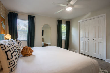 Bright modern bedroom with white bedding, black-and-white geometric pillows and a tan knot cushion, green velvet curtains, ceiling fan, arched full-length mirror, and double-door closet.