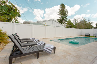 Sunny residential backyard with a rectangular pool and aqua inflatable tube, row of dark lounge chairs draped with black-and-white striped towels, beige stone patio and white privacy fence under a blue sky.