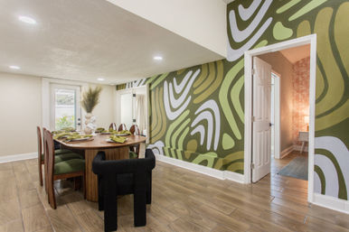 Contemporary dining room with round wooden table set for four, green upholstered chairs, playful green-and-white abstract accent wall, wood-look tile floor and open doorway to an adjacent room