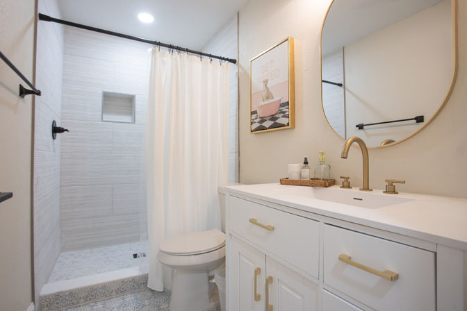 Small bright modern bathroom with white vanity and brass fixtures, round mirror, woven tray of toiletries, toilet, and walk-in shower with a white curtain over patterned tile floor.