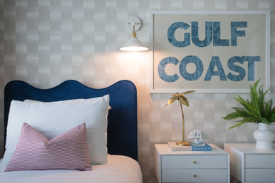 Coastal-inspired bedroom corner with a navy scalloped headboard, white pillows and red-striped lumbar pillow, white nightstands topped with a gold palm lamp, books and coral decor, a potted fern, and framed "GULF COAST" wall art under a glowing wall sconce.