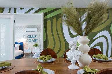 Stylish dining table set with green linen napkins, a white classical bust and a tall beige vase of pampas grass against an olive-green abstract mural, doorway revealing a Gulf Coast–themed bedroom with a blue bed
