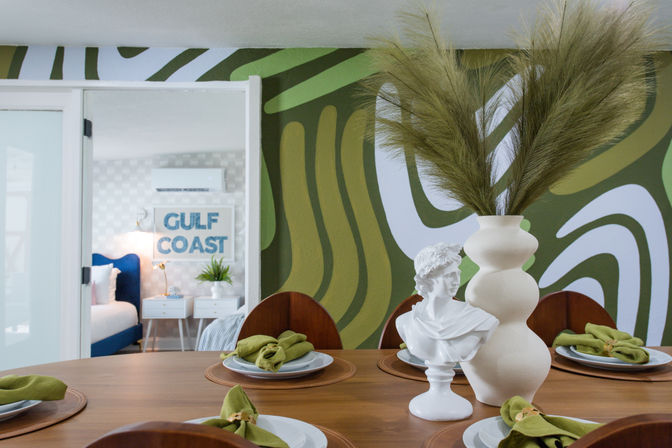 Stylish dining table set with green linen napkins, a white classical bust and a tall beige vase of pampas grass against an olive-green abstract mural, doorway revealing a Gulf Coast–themed bedroom with a blue bed