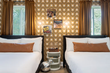 Retro-inspired twin bedroom interior with white bedding and tan leather bolster pillows, gold-and-white geometric wallpaper, brown curtains over two windows, central nightstand with a vintage record player and vinyl, illuminated wall sconces, and a disco ball on the floor.