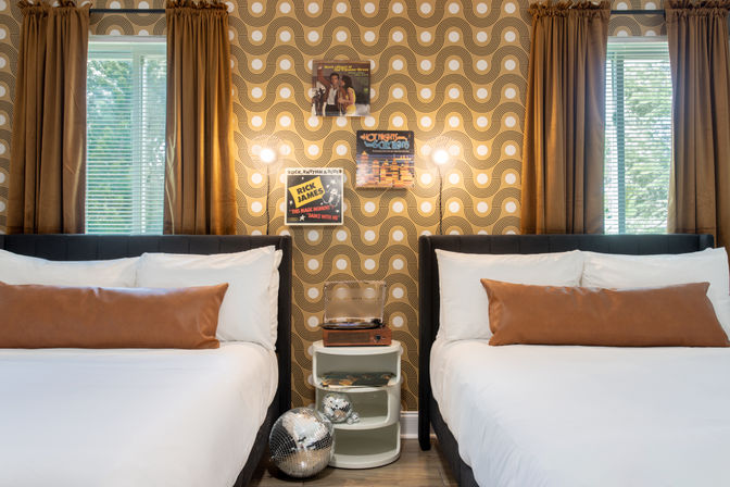 Retro-inspired twin bedroom interior with white bedding and tan leather bolster pillows, gold-and-white geometric wallpaper, brown curtains over two windows, central nightstand with a vintage record player and vinyl, illuminated wall sconces, and a disco ball on the floor.
