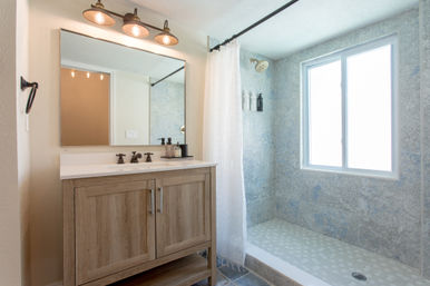 Bright modern bathroom with light wood vanity and white countertop under a three-bulb sconce, large mirror, bronze faucets, and an adjacent tiled walk-in shower with patterned floor, white curtain and a large frosted window