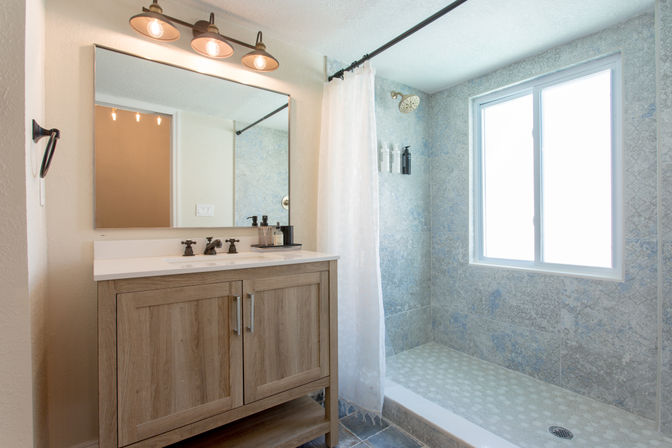 Bright modern bathroom with light wood vanity and white countertop under a three-bulb sconce, large mirror, bronze faucets, and an adjacent tiled walk-in shower with patterned floor, white curtain and a large frosted window