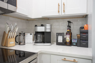 Corner coffee station on a white quartz countertop with a stainless single‑serve and drip coffee maker, French press, dark roast bag, syrup pump, knife block and pod organizer against subway tile and white cabinets with brass handles.