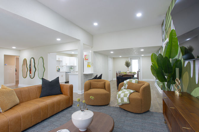 Open-concept modern living room with a caramel leather sofa, two rounded mustard armchairs with a green check throw, geometric rug and wood coffee table, large potted tropical plant and green accent wall art, with views into a bright white kitchen and dining area.