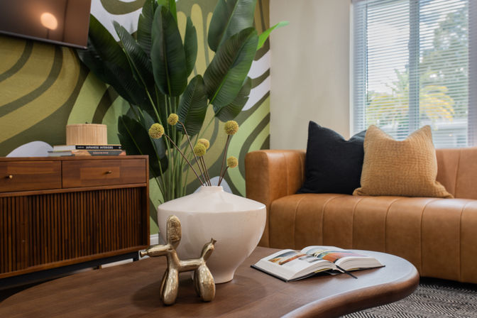 Cozy modern living room with tan leather sofa, wooden coffee table featuring a white ceramic vase of yellow billy balls and a small gold balloon-dog sculpture, tropical green mural and mid-century wooden credenza.