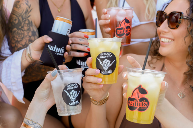 Group of friends toasting with iced summer cocktails and canned drinks at an outdoor pool party, labeled plastic cups, straws, swimwear and sunglasses.