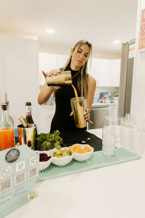 Person bartending in a modern home kitchen pouring an orange cocktail from a metal shaker into a gold mixing cup with citrus slices, mint, bottles and ice-filled glasses on the counter.