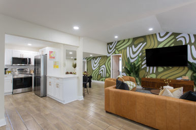 Open-concept living room and kitchen with green abstract accent wall, tan leather sectional, wall-mounted TV, mid-century wood media console, white cabinets and stainless-steel appliances on wood-look tile flooring.