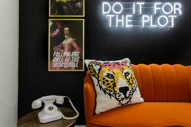 Eclectic interior corner with orange velvet sofa and tiger-printed pillow, neon wall sign reading "DO IT FOR THE PLOT", vintage white rotary phone on a side table, and framed "FOLLOW THE CALL OF THE DISCO BALL" poster on a black wall.