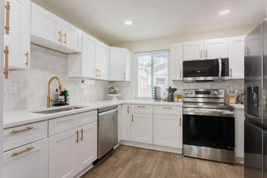 Bright modern U-shaped kitchen with white shaker cabinets, gold hardware and faucet, white tile backsplash, stainless steel appliances, wood-look flooring, and a sunny window