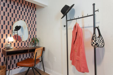 Stylish modern apartment entryway nook with pink geometric wallpaper, walnut floating desk, round mirror, lamp and leather chair, plus a black metal coat rack holding a black hat, coral corduroy coat and checkered handbag.
