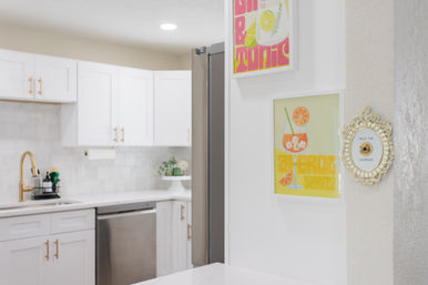 Bright modern white kitchen interior with brass faucet and hardware, stainless appliances, colorful citrus cocktail wall art and a decorative 'Press for Champagne' button.