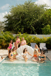 Eight women in colorful swimsuits and sunglasses enjoy a sunny backyard pool party, lounging around a white inflatable shell float with tropical palms and striped pool chairs in the background.