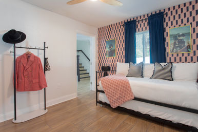 Cozy modern guest bedroom with white daybed on black metal frame, striped pillows, pink geometric accent wall, navy curtains, coral throw and a clothing rack holding a coral blazer and black hat on wood-look flooring
