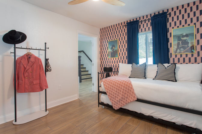 Cozy modern guest bedroom with white daybed on black metal frame, striped pillows, pink geometric accent wall, navy curtains, coral throw and a clothing rack holding a coral blazer and black hat on wood-look flooring