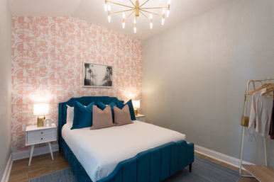 Modern bedroom interior with a teal velvet bed and white linens, teal and blush throw pillows, pink geometric accent wallpaper, white mid-century nightstands with lamps, gold starburst chandelier and open clothing rack.