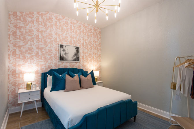 Modern bedroom interior with a teal velvet bed and white linens, teal and blush throw pillows, pink geometric accent wallpaper, white mid-century nightstands with lamps, gold starburst chandelier and open clothing rack.