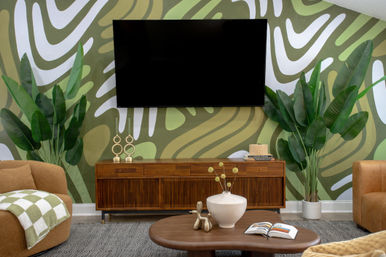 Cozy mid-century modern living room with green abstract mural, wall-mounted flat-screen TV, wooden media console, large potted tropical plants, round coffee table with vase and open book.