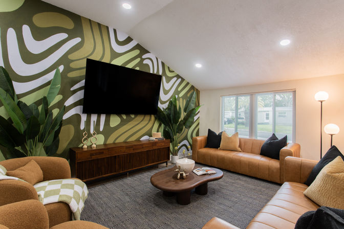 Contemporary living room with a green abstract accent wall mural, wall-mounted TV, tan leather sofas, curved armchairs, wooden media console, oval coffee table, floor lamp and large houseplants.