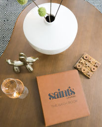 Modern lounge coffee table vignette on wood: white ceramic vase with green pom-pom stems, brass balloon-dog sculpture, wooden tic-tac-toe, rosé wine glass, and tan leather-bound book.