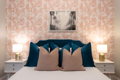 Cozy modern bedroom interior with a teal velvet headboard, layered tan and teal throw pillows, blush geometric wallpaper, white nightstands with brass lamps, and a framed black-and-white palm photo above the bed.