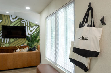 Bright modern lounge with bold green abstract accent wall and wall-mounted TV, tan leather sofa and potted plant; canvas tote bags hang on playful hand-shaped hooks beside a window with white blinds.