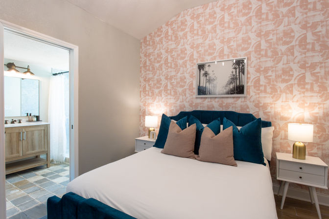 Bright modern bedroom with teal velvet headboard, white bedding and tan accent pillows against pink patterned wallpaper, flanked by white nightstands with gold table lamps and an open doorway to an ensuite bathroom with wood vanity and slate tile floor.