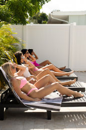 Four friends sunbathing in colorful bikinis on black lounge chairs at a sunny backyard pool deck with palm plants and a white privacy fence