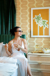 Woman in white dress spraying perfume while seated on a bed in a boutique hotel room with green velvet headboard, retro orange palm print, wavy geometric wallpaper and rattan nightstand with cocktail.