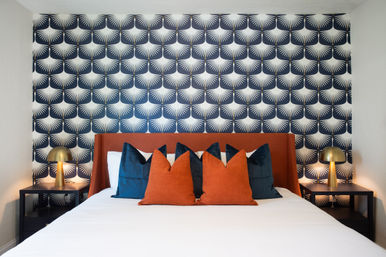 Cozy modern bedroom with navy-and-white geometric fan wallpaper, rust-orange upholstered headboard, white bedding, deep navy and rust throw pillows, and brass dome bedside lamps on black nightstands.