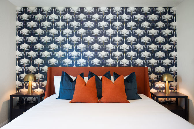 Cozy modern bedroom with navy-and-white geometric fan wallpaper, rust-orange upholstered headboard, white bedding, deep navy and rust throw pillows, and brass dome bedside lamps on black nightstands.