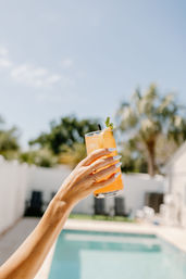 Poolside summer cocktail — hand with white manicure and rings holding a tall orange drink garnished with mint, sunny blue sky and blurred palm trees in the background.