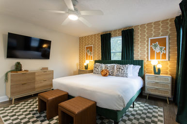 Retro-chic mid-century bedroom with king bed and green velvet headboard, geometric pillows, orange palm artwork on patterned wallpaper, wood dressers, wall-mounted TV, ceiling fan and checkered rug