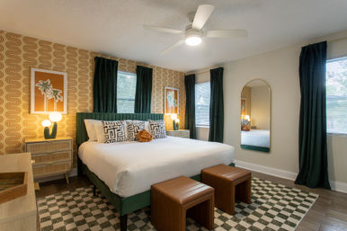 Tropical-meets-retro bedroom interior with king bed and green velvet headboard, geometric pillows, wavy orange wallpaper, dark green curtains, ceiling fan, bedside lamps, leather ottomans and checkered area rug.