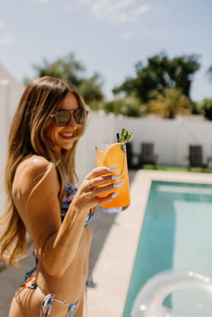 Smiling woman in sunglasses and a colorful bikini holding a bright orange, mint-garnished cocktail poolside on a sunny summer day.