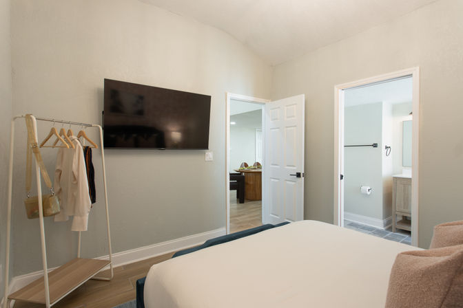 Modern minimalist bedroom interior with white bedding, wall-mounted flat-screen TV, freestanding clothing rack with a few garments, and open doors revealing a bathroom and adjoining room.