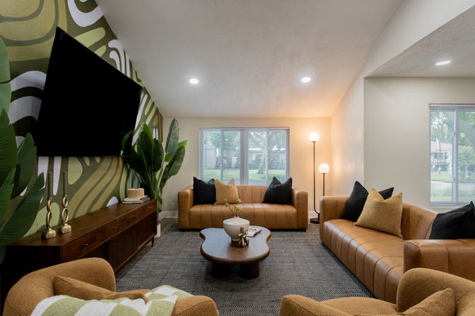 Cozy retro-meets-modern living room with tan leather sofas and armchairs, patterned rug, sculptural wood coffee table, green botanical accent wall with mounted flat-screen TV, tall indoor plants, floor lamp, and large windows looking out to a suburban yard.