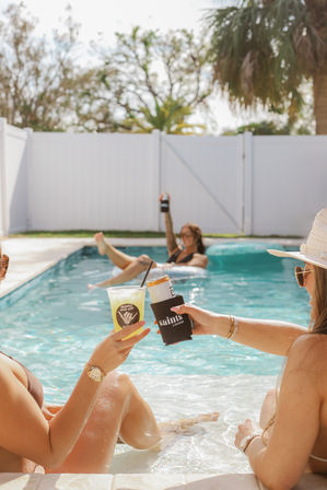 Two friends relaxing at a sunny backyard pool, toes in the water and clinking cold summer drinks while a third person floats on an inner tube