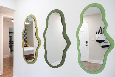 Playful wavy green-framed mirrors on a white entryway wall reflecting a bedroom, front door, light wood floors and hanging tote bags.