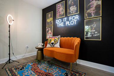 Stylish studio corner with an orange scalloped loveseat and tiger pillow under a 'DO IT FOR THE PLOT' neon sign, blue tiger rug, ring light and framed art on a black accent wall.