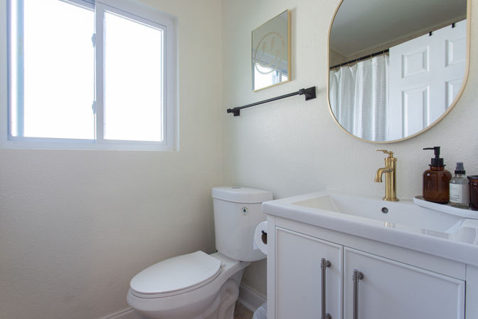 Bright small modern bathroom with white toilet and vanity, gold faucet, round mirror, amber soap dispensers, towel bar and a window letting in natural light.