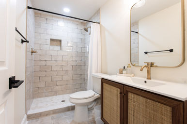 Bright spa-like modern bathroom with a walk-in tiled shower and white curtain, brass showerhead and faucet, rattan-front wooden vanity with mirror, and a white toilet.