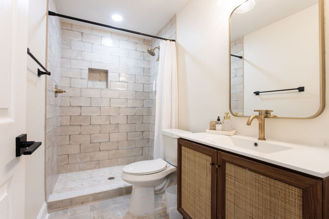 Bright spa-like modern bathroom with a walk-in tiled shower and white curtain, brass showerhead and faucet, rattan-front wooden vanity with mirror, and a white toilet.