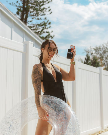 Smiling woman in a black one-piece swimsuit and sunglasses holding a canned drink in a koozie and a clear glitter swim ring by a white backyard fence on a sunny summer day.